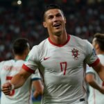 Cristiano Ronaldo celebrates scoring the decisive goal for Portugal against France in the UEFA Nations League semi-final at Stuttgart's MHPArena, with Portuguese players in white and red kits embracing under floodlights.