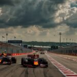 A dynamic Formula 1 race scene at Circuit of the Americas, featuring two high-speed cars driven by Max Verstappen and Lando Norris racing up the iconic 20-degree climb out of Turn 1, with a packed stadium crowd and a dramatic Texas sky in the background.